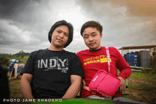a man wearing a black indy shirt sits next to another man in a red supreme shirt