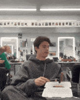 a man in a grey hoodie is eating a hot dog from a styrofoam box