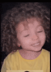 a little girl with curly hair wearing a yellow shirt is smiling .