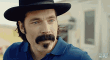 a man with a mustache wearing a hat and a blue shirt with the letters hw on it