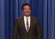 a man in a suit and tie holds his hand to his chest in front of a blue curtain