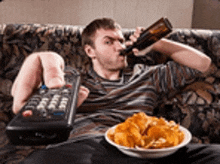 a man is laying on a couch drinking beer and eating chips .