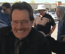a man wearing glasses and a hat smiles in front of a group of people