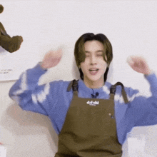 a young man wearing an apron and a blue sweater is sitting on a chair with his arms outstretched .