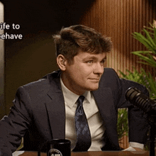 a man in a suit and tie is sitting at a table in front of a microphone with the words life to behave above him
