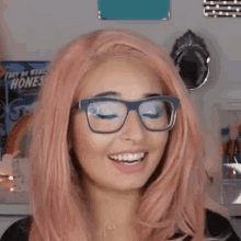 a woman with pink hair and glasses is smiling in front of a rainbow poster .