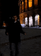 a man in a black jacket stands in front of a large building at night