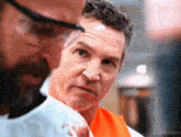a man with a beard wearing a white shirt and an orange vest looks at another man