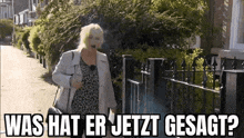 a woman is walking down a street in front of a fence with the words `` was hat er jetzt gesagt '' .