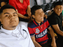 a man wearing a red and blue england shirt sits next to another man