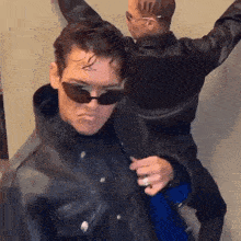 a man in a leather jacket and sunglasses is standing next to another man .