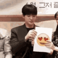 a man in a suit is holding a piece of paper with an emoji face with hearts in his eyes .