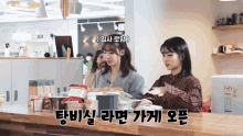 two women are standing behind a counter with korean writing on it .