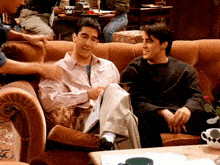 two men are sitting on a couch talking and one of them is holding a piece of paper