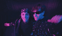 a man wearing sunglasses and a bandana stands next to another man wearing sunglasses