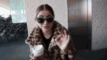 a woman wearing sunglasses and a fur coat is holding a cup of coffee and giving an okay sign .
