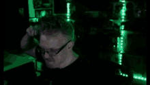 a man with glasses is sitting in a dark room with green lights on the wall