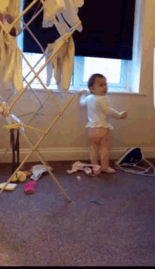 a baby standing in front of a window with clothes hanging on a drying rack