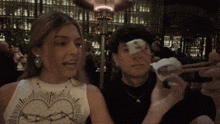 a woman wearing a white tank top with a heart on it looks at a man holding a piece of paper