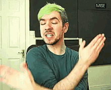 a man with green hair and a beard is clapping his hands in front of a sign that says '  mise '
