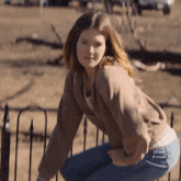 a woman in a brown sweatshirt and blue jeans is squatting on a fence