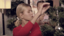 a woman in a red shirt is decorating a christmas tree with a red ornament .