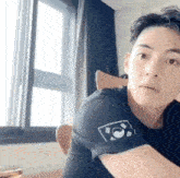 a young man is sitting in front of a window wearing a black shirt with a logo on the sleeve .