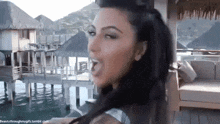 a woman with long black hair is standing in front of a body of water with her tongue out .