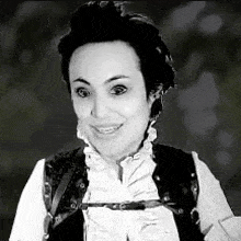 a woman is smiling in a black and white photo . she is wearing a white shirt and a black vest .
