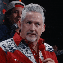 a man in a red and white shirt stands in front of a microphone with another man in a white hat behind him