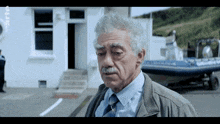 a man with gray hair and a mustache is standing in front of a white building with a boat parked in front of it