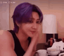 a man with purple hair is sitting at a table with a cup of coffee and a lamp .