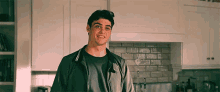 a young man in a jacket is standing in a kitchen smiling .