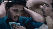 a man in a blue shirt is looking at a cell phone while another man looks on .