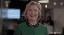 hillary clinton is wearing a green jacket and smiling in a room .