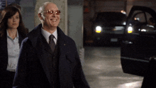 a man wearing sunglasses and a suit smiles while a woman walks behind him