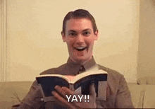 a man is sitting on a couch holding a book and saying yay .