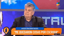 a man in a suit and polka dot shirt is sitting in front of a sign that says " me quedaran cosas por escribir "