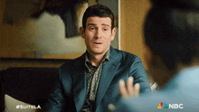 a man in a suit is sitting on a couch talking to a woman with the nbc logo in the corner