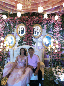 a woman in a pink dress sits next to a man in a pink shirt in front of a floral wall with the number 18 on it