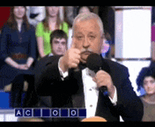 a man in a tuxedo is speaking into a microphone in front of a crowd of people .