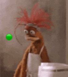 a muppet with a red hair and a white apron is standing in front of a green ball .