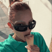 a woman wearing sunglasses and a green shirt is biting into an ice cream cone