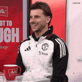 a man wearing a manchester united jacket is smiling in front of a canirove sign