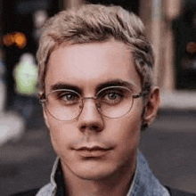 a young man wearing glasses and a denim jacket is standing on a street .