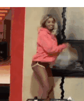 a woman in a pink hoodie and shorts is dancing in front of a fireplace in a living room .