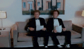 two men in tuxedos sit on a couch with papers