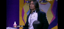 a man with long hair is sitting in front of a painting of a man 's face .