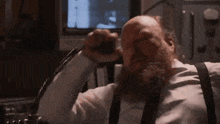 a bald man with a beard and suspenders is talking on a phone