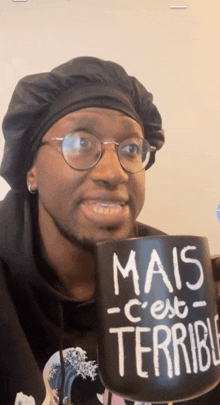 a man wearing glasses and a black hat holds a black mug that says mais c'est terrible